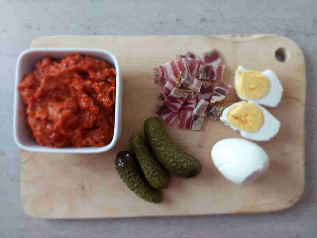 An example of ajvar being served with bacon, pickles, and boiled eggs